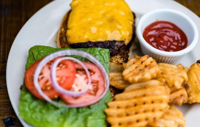 A Cafe Gecko Eastside classic: a cheeseburger with cheddar, lettuce, tomato, and onion, served with waffle fries and a small cup of ketchup on a white plate.