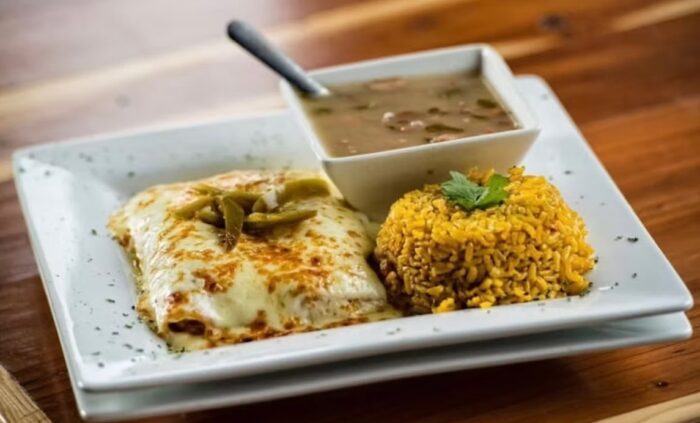 A plate with a cheesy enchilada topped with jalapeños, a serving of yellow rice garnished with parsley, and a bowl of beans with a spoon, all beautifully arranged on a wooden table at Cafe Gecko Eastside.