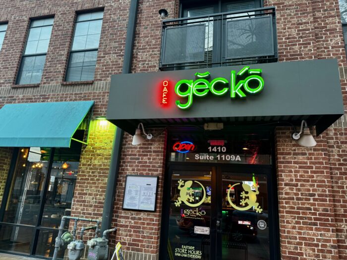 A brick building with a green neon Cafe Gecko Eastside sign above a glass door. There are menus posted outside, a blue-green awning next door, and decorative gecko decals on the entrance.