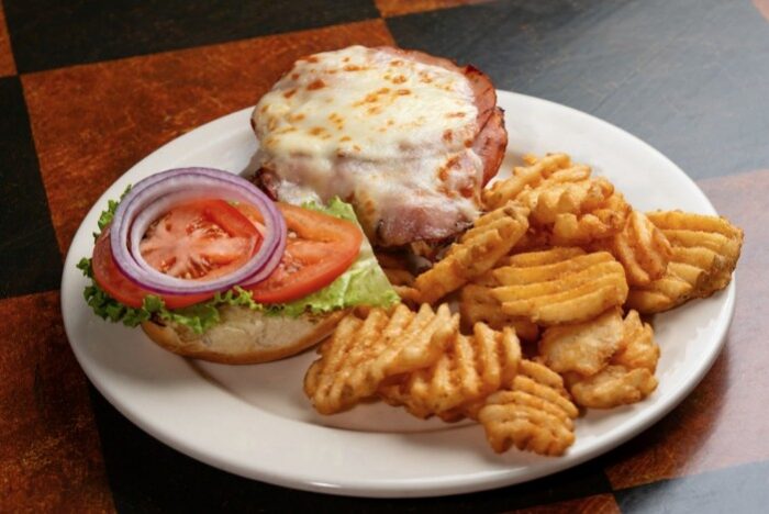 A plate with a sandwich topped with melted cheese, ham, lettuce, tomato, and onion, served open-faced alongside crisscrossed waffle fries—a favorite at Cafe Gecko Eastside.