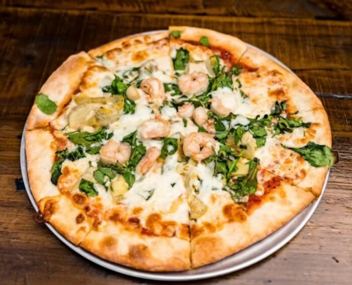 A pizza from Cafe Gecko Eastside topped with shrimp, spinach, artichoke hearts, and melted cheese sits on a metal tray atop a wooden table.