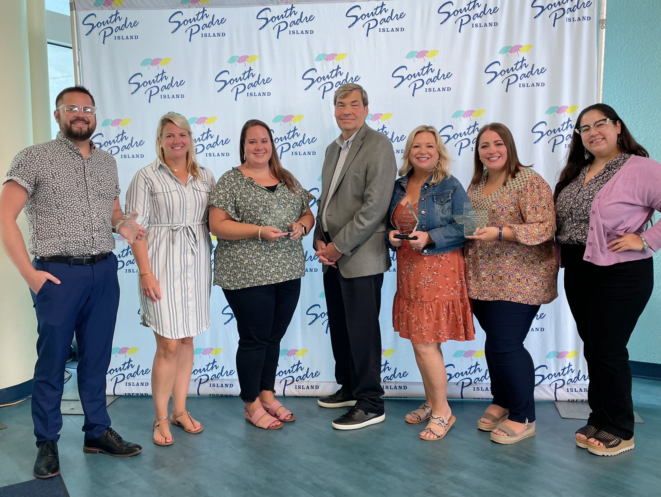 Visit Plano staff holding Texas Destination Awards in front of a South Padre Island backdrop