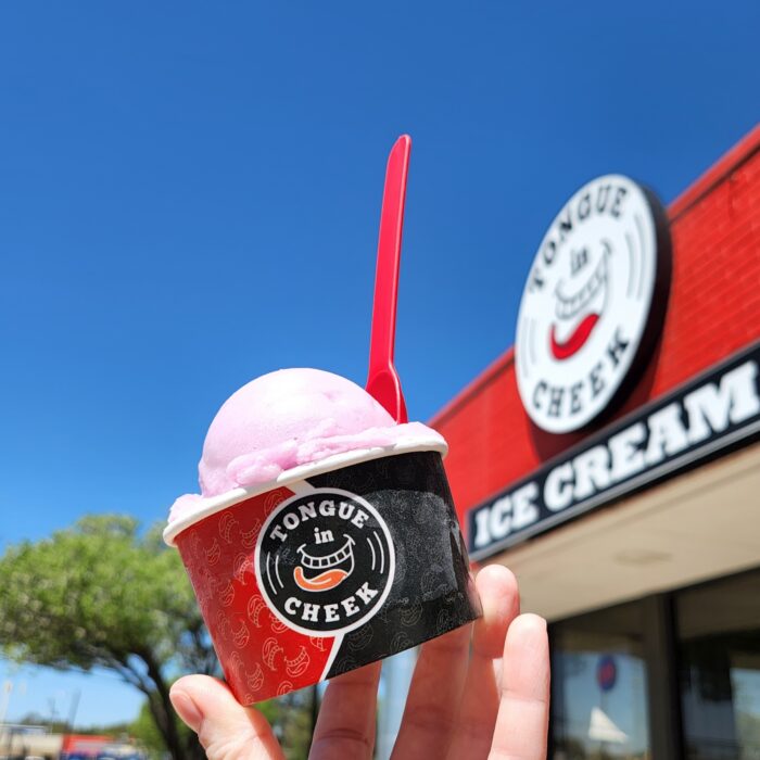 A hand holds a cup of pink ice cream with a red spoon in front of a building with a sign that reads Tongue in Cheek Ice Cream under a clear blue sky.