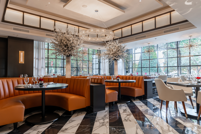 Elegant Truluck's restaurant interior with orange curved booths, white chairs, black tables, marble floors, large windows, chandeliers, and decorative trees with white blossoms, all creating a bright and upscale dining atmosphere.