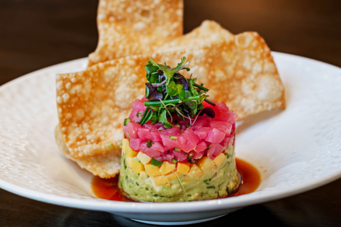 A stack of diced avocado, mango, and tuna tartare topped with microgreens is artfully served at Truluck’s, accompanied by crispy wonton chips on a white plate with soy sauce.
