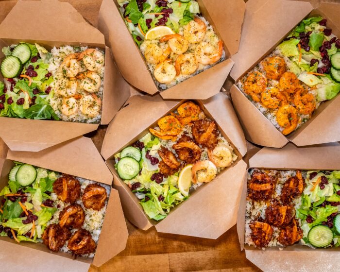 Seven brown takeout boxes filled with rice, grilled shrimp, fresh green salad with cucumber slices, cranberries, and lemon wedges, arranged on a wooden surface.