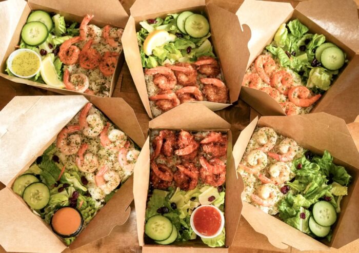 Six takeout boxes filled with shrimp, rice, and green salads with cucumbers, tomatoes, and lemon wedges, arranged in a circle. Two boxes include orange and red dipping sauces in small cups.