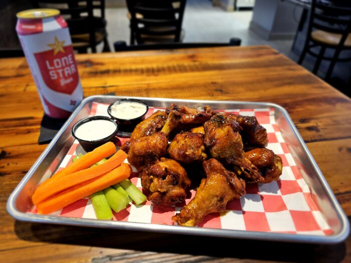 A tray of glazed chicken wings with celery and carrot sticks, two cups of ranch dip, and a can of Lone Star beer on a wooden table at Dirty Birds Sports Bar & Grill.