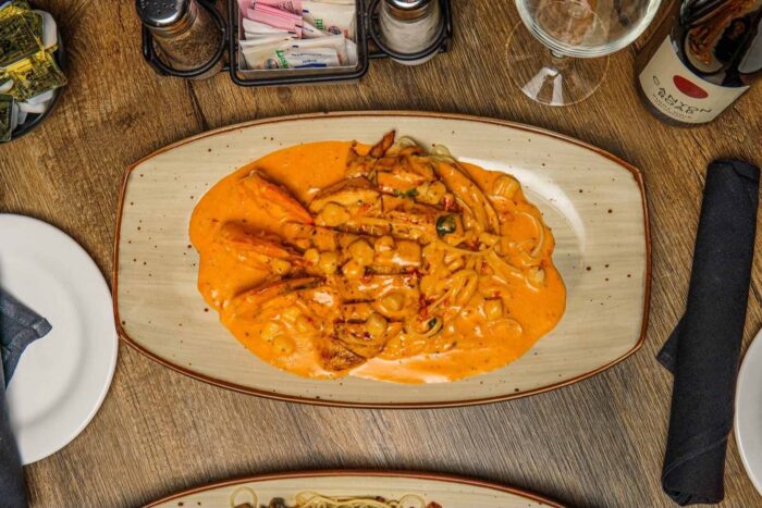 A rectangular plate with grilled chicken breast topped with a creamy orange sauce and sliced vegetables, placed on a wooden table set with a wine glass, napkins, and condiments.