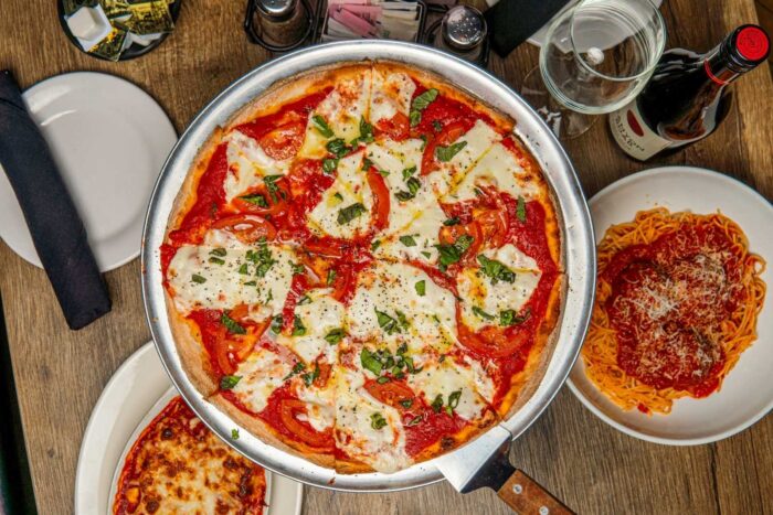 A table with a large margherita pizza topped with mozzarella, tomatoes, and basil, surrounded by plates of pasta with red sauce, a baked dish, a glass of water, and a bottle of red wine.