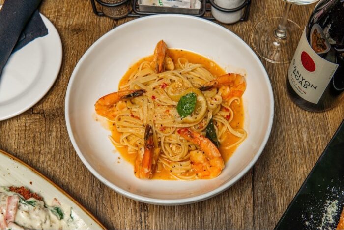 A bowl of linguine pasta with shrimp in a tomato-based sauce, garnished with basil, is served on a wooden table with a bottle of red wine and a plate of food partially visible beside it.
