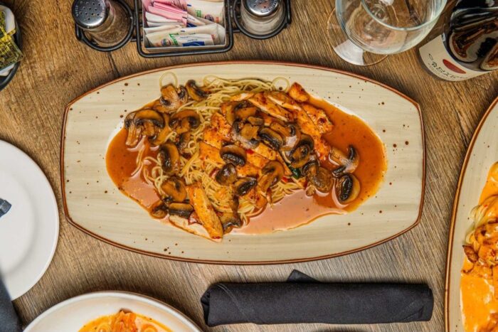 A plate of spaghetti topped with sliced grilled chicken, mushrooms, and brown sauce sits on a wooden table set with a napkin, utensils, a wine glass, and condiments.