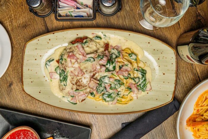 A plate of creamy pasta topped with ham, spinach, and melted cheese, served on an oval dish. A glass of wine, condiments, and another plate of pasta are visible on the wooden table.