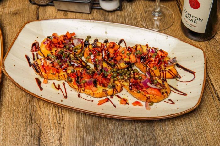 A plate of bruschetta topped with diced tomatoes, red onions, capers, herbs, and a drizzle of balsamic glaze, served on a rustic wooden table with a bottle of red wine in the background.