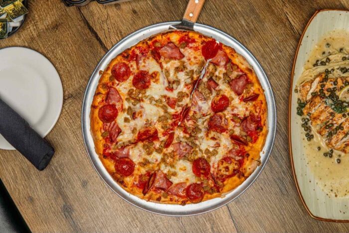 A round meat pizza with pepperoni, sausage, and ham sits on a metal tray with a pizza cutter. Nearby are a plate with a black napkin and a rectangular dish with food on a wooden table.