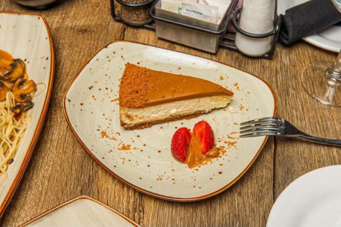A slice of caramel-topped cheesecake on a square plate, garnished with two sliced strawberries and a dollop of caramel, with a fork beside it on a wooden table.