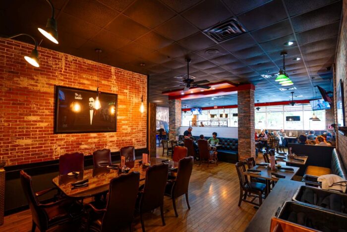 A warmly lit restaurant with brick walls, a large framed photo, wooden tables and chairs, people dining, and TVs mounted on the walls. The atmosphere appears cozy and inviting.