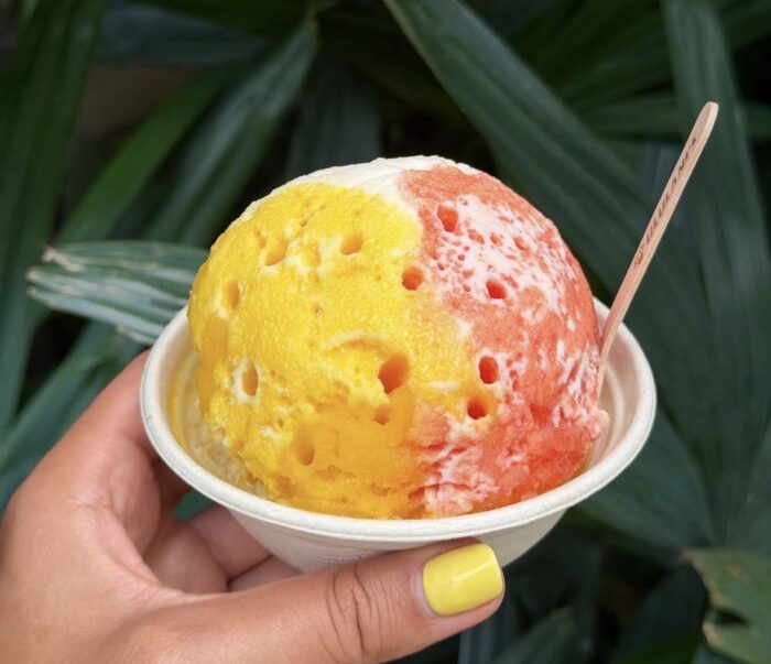 A hand with yellow-painted nails holds a bowl of colorful Ululani's Hawaiian Shave Ice in yellow, orange, and red sections, with a wooden spoon, set against a background of green leaves.