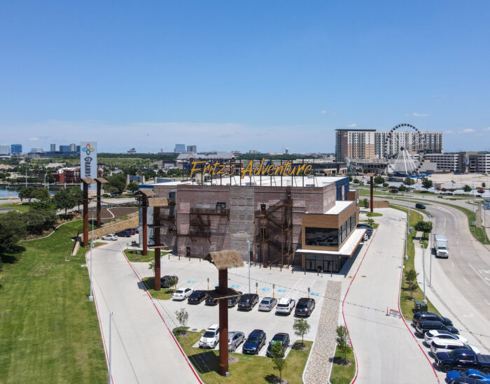 A large adventure park building with a sign reading “Fritz’s Adventure” sits near a winding road and parking lot, with a grassy area, city buildings, and a Ferris wheel visible in the background under a clear blue sky.