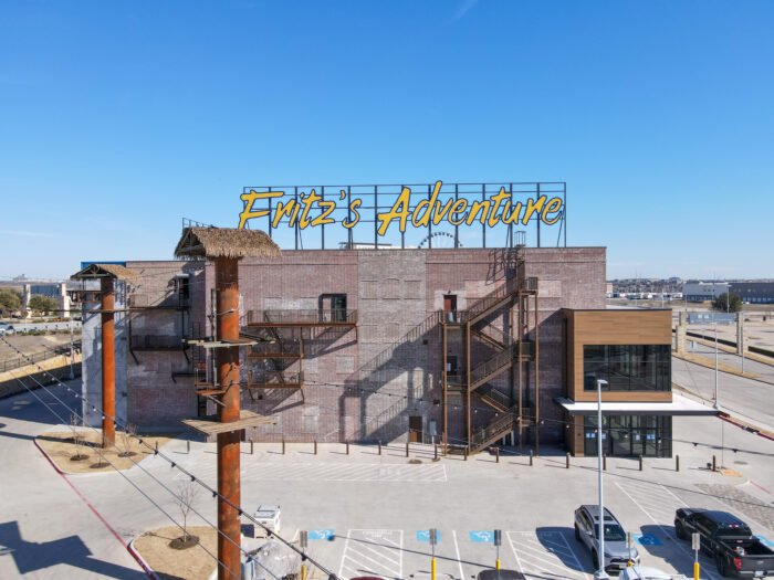 A large brick building with a yellow sign reading Fritzs Adventure on the roof, featuring metal staircases, climbing structures, and an empty parking lot in front under a clear blue sky.