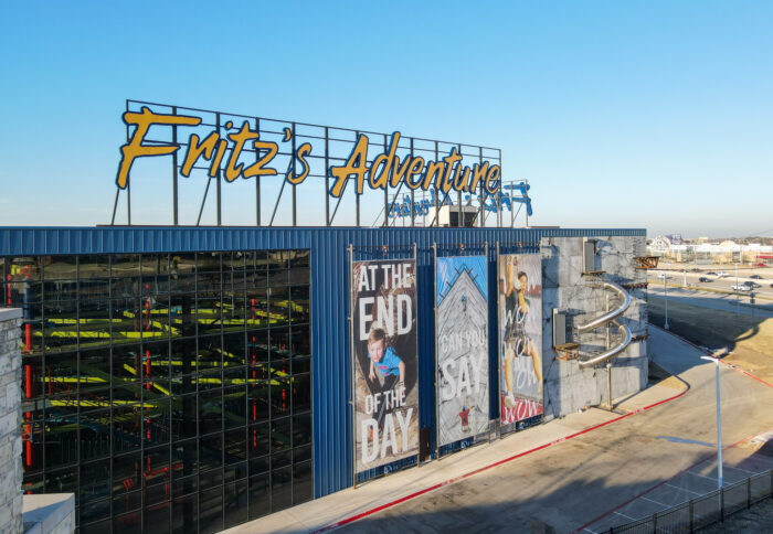 Large blue building with a yellow sign reading Fritzs Adventure. Colorful banners on the side show people engaged in activities. A winding metal slide extends from the upper floor to the ground. Blue sky and parking lot visible.