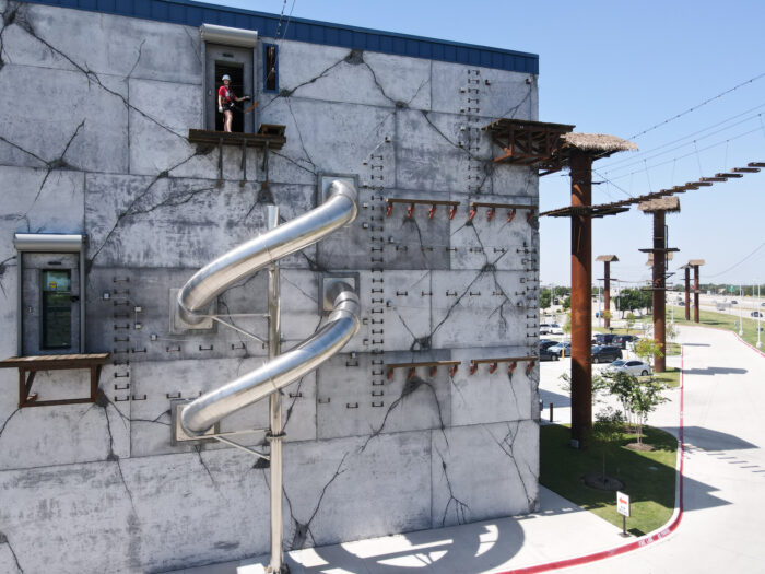 A person stands on a platform attached to a tall, cracked concrete wall with a metal spiral slide and climbing holds. Rope bridges and obstacle course elements are visible to the right. It’s a sunny day.