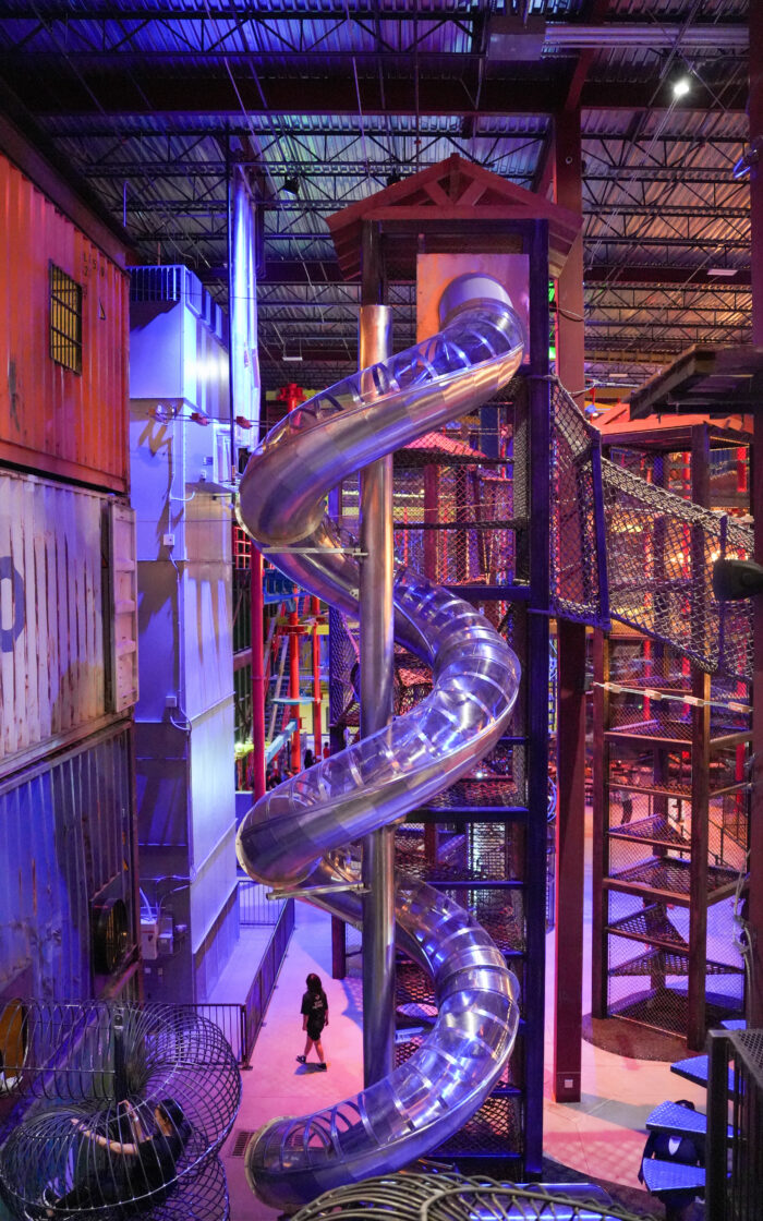 A tall, silver spiral slide winds down a metal tower inside an indoor playground with climbing structures and netting. A person walks below, and the space is lit with blue and purple lights.