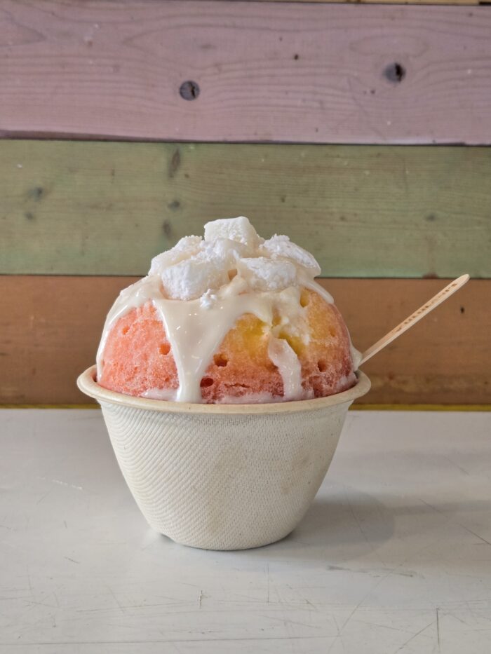 A bowl of colorful Ululani's Hawaiian Shave Ice with white syrup and coconut chunks on top, served in a textured cup with a wooden spoon. The background features pastel-colored wooden planks.