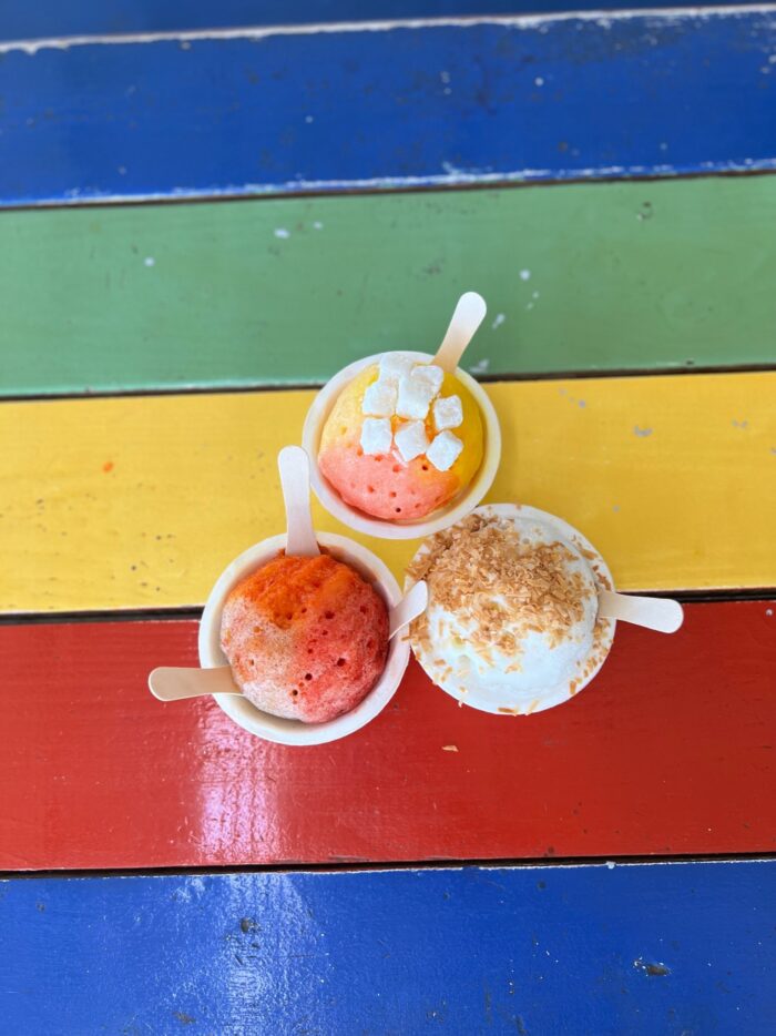 Three cups of colorful Ululani's Hawaiian Shave Ice with various toppings and wooden spoons sit on a vibrant table painted with green, yellow, blue, and red stripes.