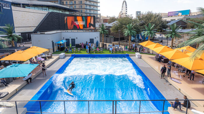 A person surfs on an artificial wave pool at PopStroke, surrounded by people, palm trees, and colorful umbrellas in an outdoor recreational area with shops and a Ferris wheel in the background.