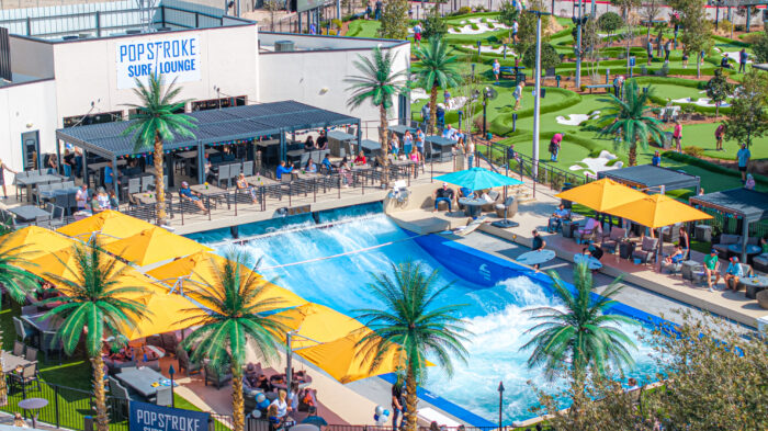 A vibrant outdoor entertainment area at PopStroke features a mini-golf course, palm trees, shaded seating, and a blue artificial wave pool at the Surf Lounge, with people enjoying various activities.