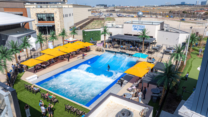 Aerial view of an outdoor surf simulator at PopStroke with people watching under yellow umbrellas, artificial palm trees, surrounding seating, and a nearby bar and lounge in a commercial area.