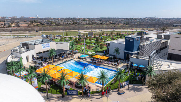 Aerial view of a recreational complex featuring an artificial wave pool with surfers, surrounded by palm trees, orange umbrellas, and people. Nearby are PopStroke mini-golf courses, buildings, grassy areas, and a cityscape in the background.