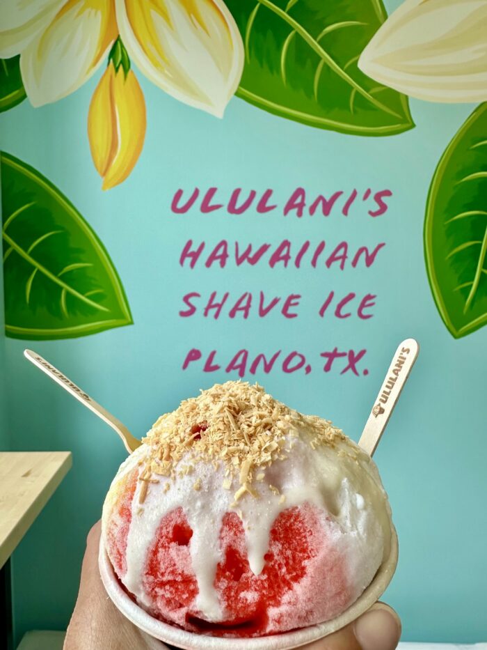 A hand holds a bowl of Ululani's Hawaiian Shave Ice topped with coconut flakes, set against a vibrant mural of green leaves and the words “ULULANI’S HAWAIIAN SHAVE ICE PLANO, TX.” in bold pink letters.