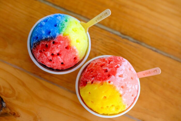 Two bowls of Ululani's Hawaiian Shave Ice with bright red, yellow, and blue flavors in sections, served with wooden spoons on a wooden surface.