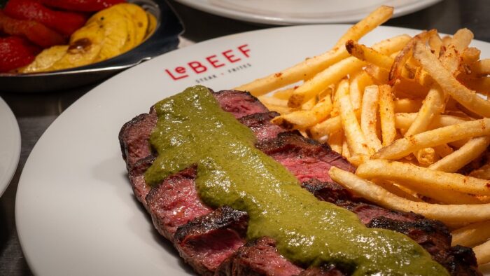 A plate with grilled steak topped with green sauce and a generous serving of French fries, served on a white plate with “LeBEEF” branding. In the background, there is a side dish of grilled vegetables.