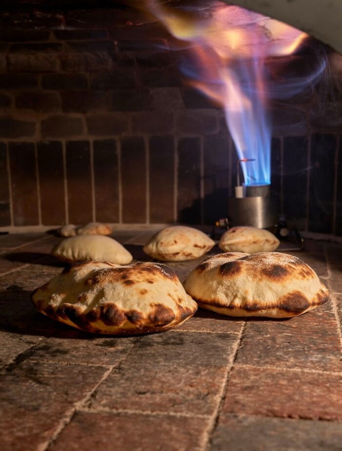 Pita breads puff up as they bake on the hot stone floor of a brick oven, with a blue flame burner in the background against the brick wall—evoking classic Harissa Mediterranean flavors.