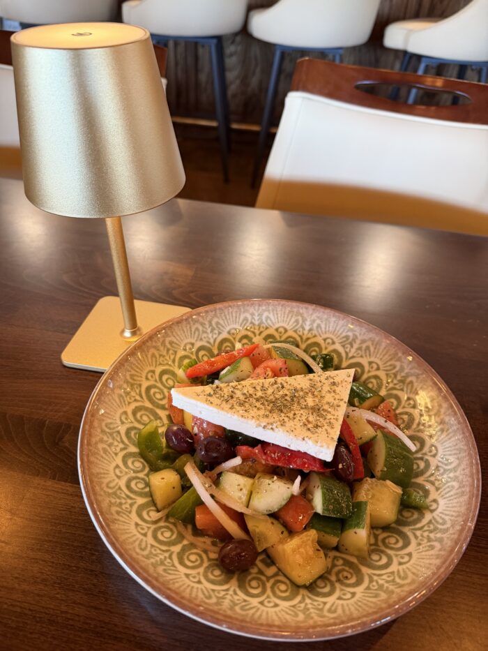 A bowl of Greek salad with tomatoes, cucumber, olives, onions, and a slice of feta cheese sits on a wooden table beside a small, gold-colored lamp. Harissa adds a Mediterranean kick. White chairs are visible in the background.