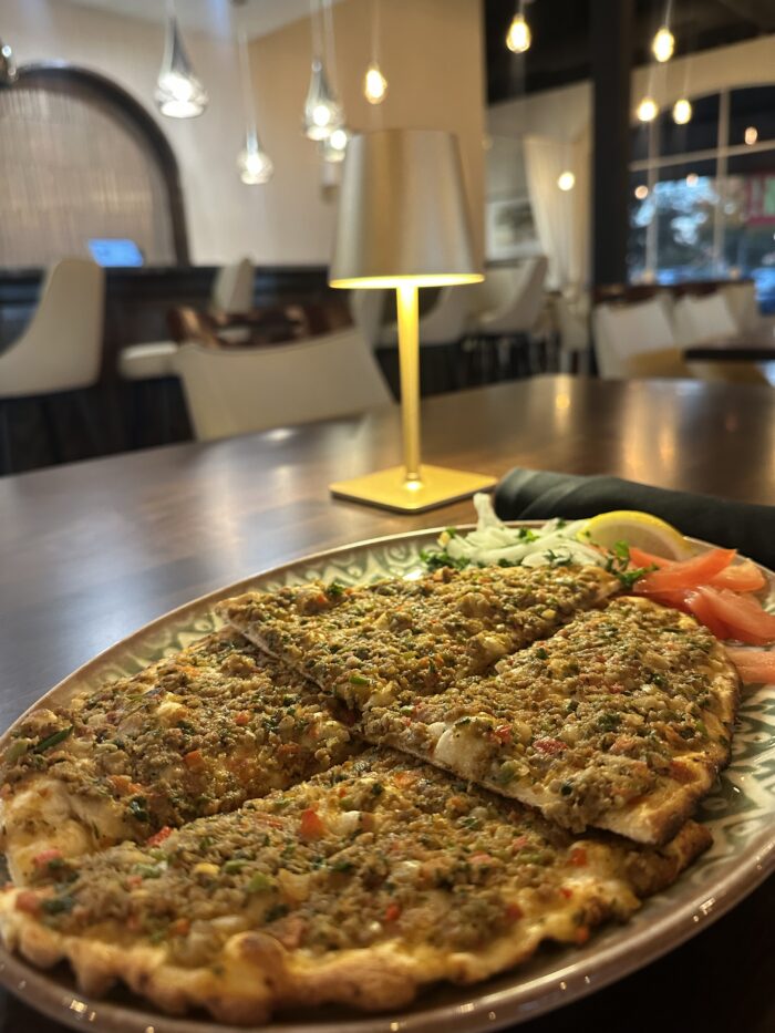 A plate of sliced flatbread topped with finely chopped meat and vegetables sits on a wooden table in a modern, softly lit Harissa Mediterranean restaurant, with a small lamp and fresh salad garnish beside the bread.