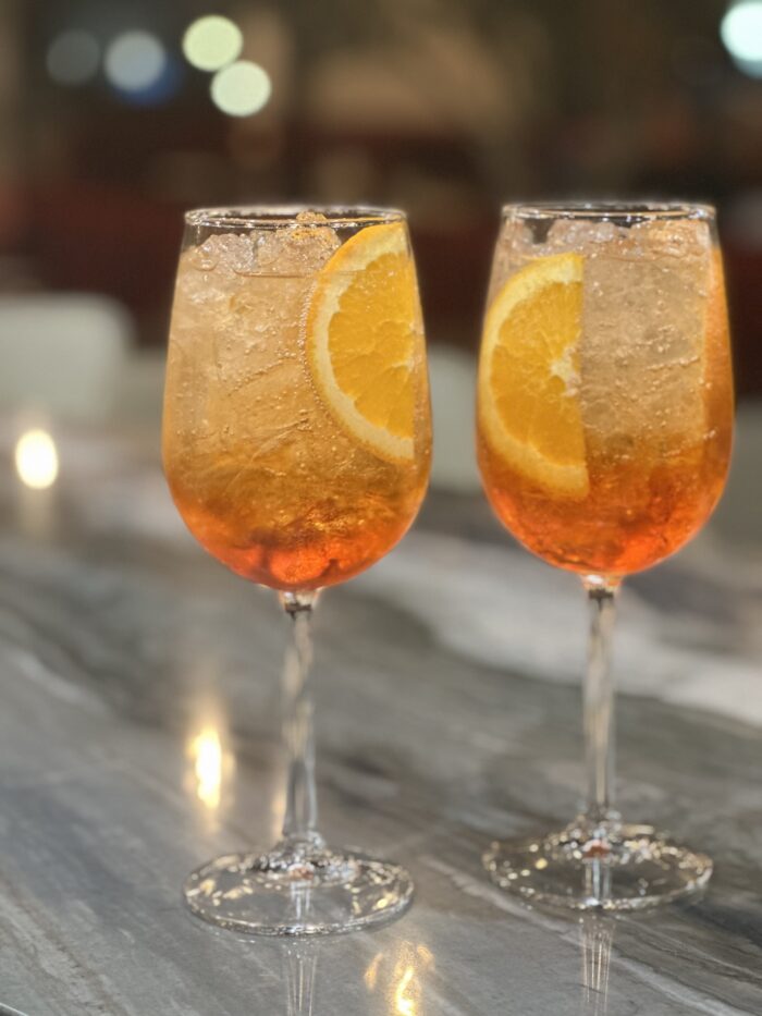 Two stemmed glasses filled with an orange-colored Harissa Mediterranean cocktail, ice cubes, and garnished with orange slices sit on a marble surface with a softly blurred background.