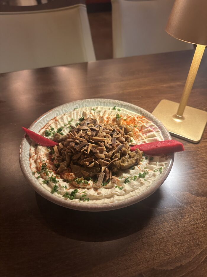 A bowl of Harissa-spiced Middle Eastern mezze topped with sautéed eggplant, slivered almonds, chopped parsley, and pickled turnip sits on a wooden table next to a small gold lamp, evoking rich Mediterranean flavors.