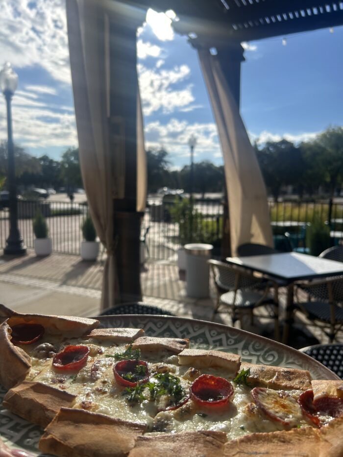 A close-up of a Harissa Mediterranean flatbread pizza topped with pepperoni and herbs on a patterned plate, outdoors on a sunny patio with empty tables, chairs, and trees in the background.
