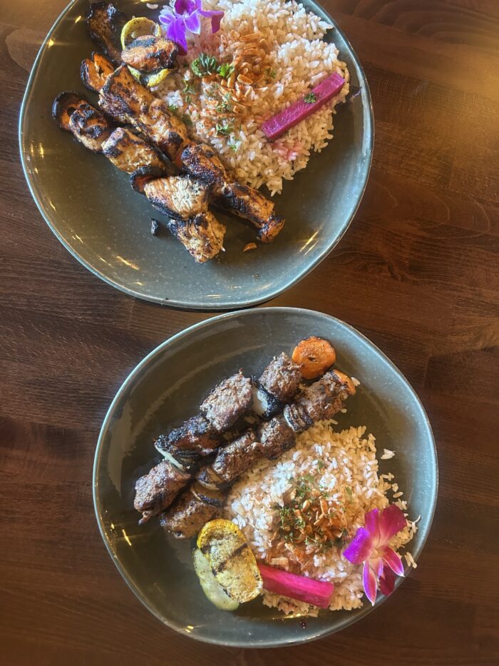 Two green plates each with skewered grilled meat, white rice topped with fried onions, grilled vegetables, and pink pickled garnish, served on a wooden table and decorated with edible flowers and a touch of Harissa Mediterranean flavor.
