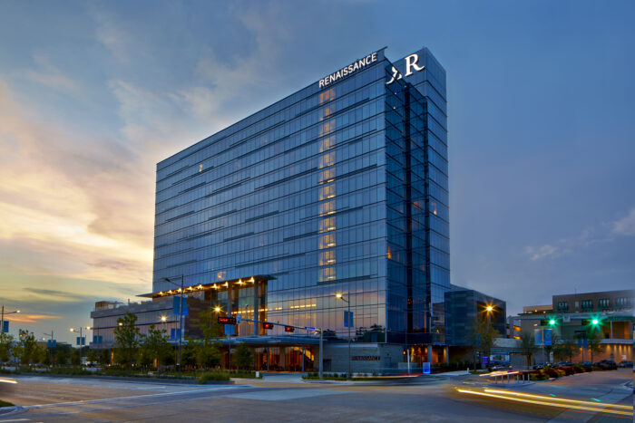 A modern, glass-covered hotel labeled Renaissance stands tall at dusk in Gather in Plano, with warm lights illuminating its entrance and cars passing by on the street below.