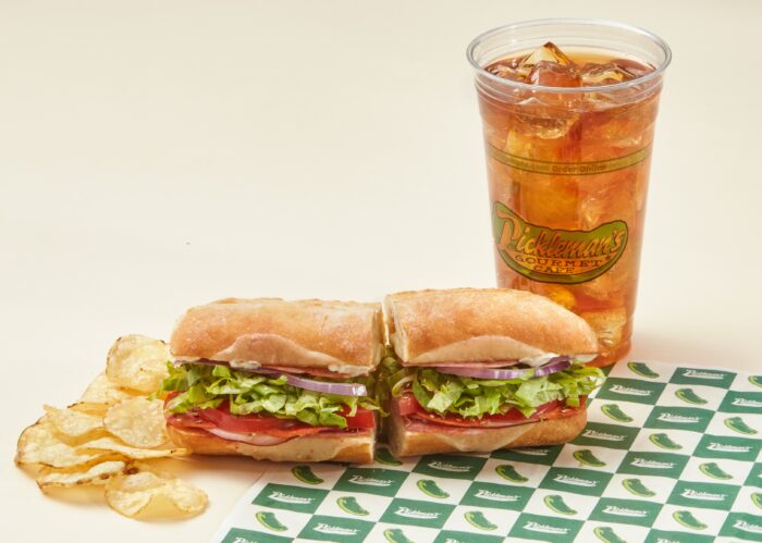 A sub sandwich with lettuce, tomato, onion, and deli meat sits on branded paper next to a pile of potato chips and a large cup of iced tea with the Pickleman’s Gourmet Cafe logo.