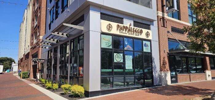 A corner view of PappaLecce Espresso Gelato Studio, reminiscent of Pappalecco, a café in a modern brick building with large glass windows, signage, and small shrubs along the sidewalk on a sunny day.