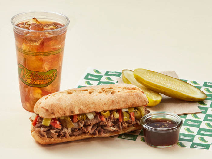 A toasted sandwich filled with sliced beef, peppers, and onions sits on sandwich paper next to a cup of iced tea, two pickle spears, a cup of au jus, and a napkin.