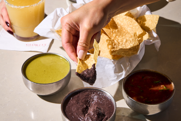A hand dips a tortilla chip into black bean dip, with bowls of green salsa and red salsa nearby, alongside a basket of chips and a glass of yellow beverage on a table.