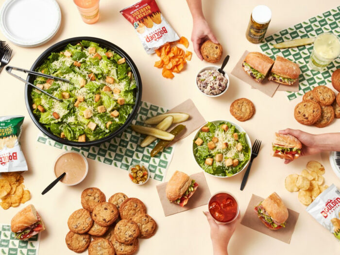 A table set with a large Caesar salad, sandwiches, potato chips, pickles, cookies, dipping sauces, and hands reaching for food, creating a casual meal or gathering atmosphere.
