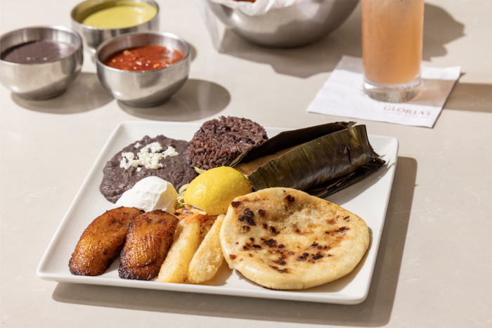A white plate with Salvadoran food, including a pupusa, plantains, yuca, tamale in banana leaf, rice, black beans with cheese, sour cream, and a lemon wedge. Sauces and a drink are in the background.